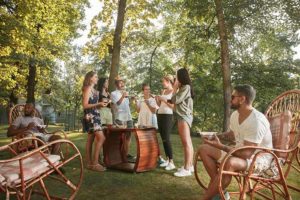 Group of happy friends eating and drinking beers at barbecue dinner on sunset time. Having meal together outdoor in a forest glade. Celebrating and relaxing. Summer lifestyle, food, friendship concept.